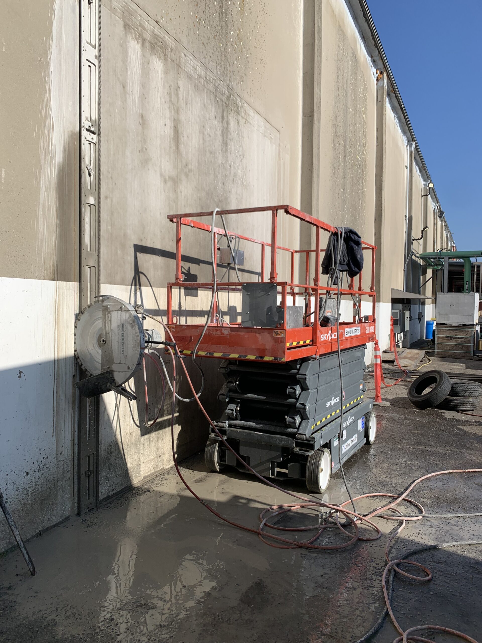 Worker lift equipment is positioned against a warehouse wall being cleaned. Water splashes accumulate on the ground, showcasing maintenance in progress.